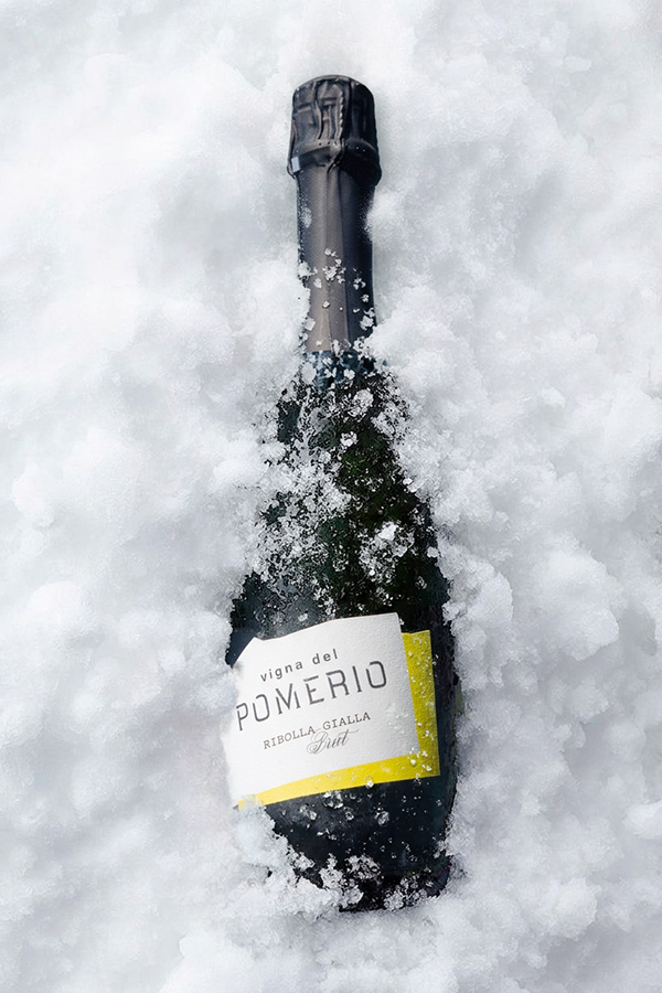 Bottiglia di vino Ribolla Gialla spumante Brut Millesimato Pomerio in primo piano, parzialmente coperta da neve fresca.