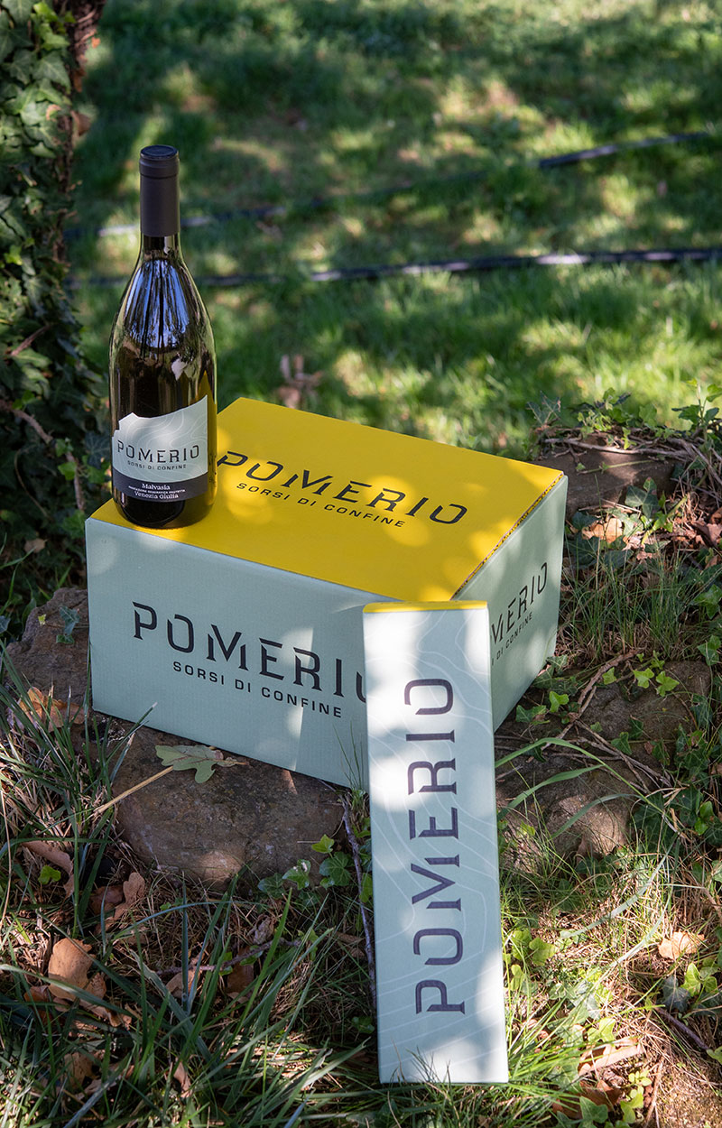 Confezioni da una e da sei bottiglie di vino bianco Pomerio IGT Venezia Giulia, con una bottiglia appoggiata sulla scatola all'ombra di un albero in una giornata estiva.