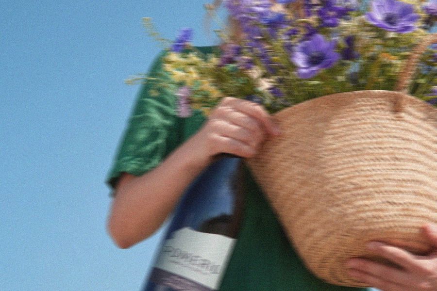 Donna in un paesaggio estivo soleggiato che tiene in mano un cesto di fiori e una bottiglia di vino bianco Pomerio IGT Venezia Giulia.
